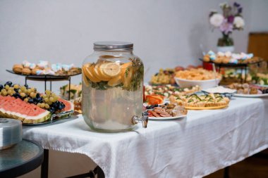 Güzel düzenlenmiş bir tatlı masasında, beyaz bir masa örtüsü üzerinde çeşitli renkli meyveler, hamur işleri ve atıştırmalıklarla çevrili limon suyuyla dolu büyük bir cam şeker kutusu bulunur.