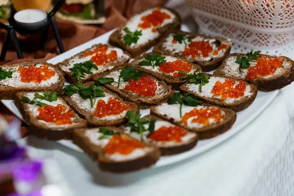 Yakın plan çekimde üzerine beyaz krem peynir, canlı kırmızı havyar ve taze maydanoz serpiştirilmiş koyu çavdar ekmeği kanepeleri sergilenir. Beyaz bir tabakta lüks bir aperatif düzenleme.