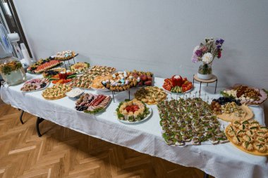 Uzun beyaz masa örtüsü renkli mezeler, taze meyveler, sebzeler, peynirler ve bir kutlama için parti dekorasyonlarıyla geniş bir ziyafet sergiliyor.