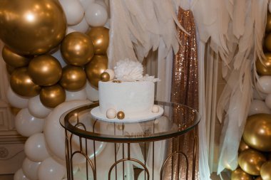İnce altın aksanlı güzel buzlu beyaz bir pasta cam bir masanın üzerinde duruyor, beyaz tüyler ve altın balonlardan oluşan bayramlık bir zemin üzerinde, bir kutlamayı simgeliyor.