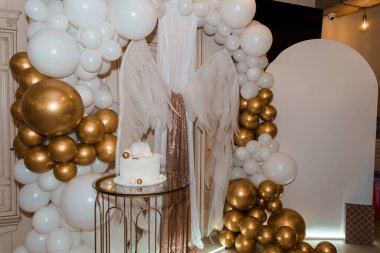 Çok şık bir ilk doğum günü partisinde muhteşem beyaz ve altın rengi bir balon, melek kanatlı bir düzeneği küçük bir pastayla çerçeveleyerek neşeli bir kutlama havası yakalıyor.