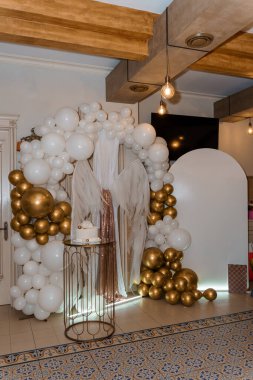 Büyüleyici melek temalı bir parti düzeninde büyük beyaz ve altın renkli bir balon, melek kanadıyla süslenmiş bir pasta masasını çerçeveleyerek neşeli bir ortam yaratıyor.