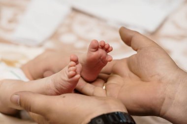 Sevgi dolu bir baba, yeni doğmuş bebeğini nazikçe elinde tutar. Küçük ayakları, yeni ebeveynliği, aile bağlarını ve yumuşak bej arkaplanlı şefkati simgeler.