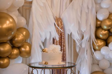 Güzel, beyaz, buzlu bir doğum günü pastası, beyaz balonlar ve akıcı kumaşlarla çerçevelenmiş altın bir masanın üzerine kurulur. Kutlama görüntüsü için ideal.