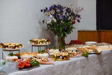 Güzel düzenlenmiş bir açık büfe yemek masasında bol bol kek, peynir, meyve ve mezeler sergileniyor. Çiçeklerle süslenmiş vazoda.
