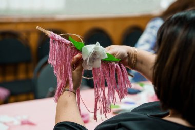 Bir kişinin elleri, muhtemelen bir atölye ya da DIY sanat etkinliği sırasında masada beyaz bir çiçekle pembe püsküllü bir süsleme yaparken gösteriliyor.