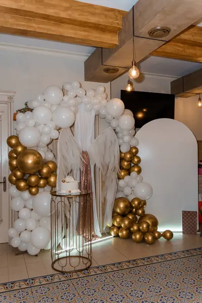 Büyüleyici melek temalı bir parti düzeninde büyük beyaz ve altın renkli bir balon, melek kanadıyla süslenmiş bir pasta masasını çerçeveleyerek neşeli bir ortam yaratıyor.