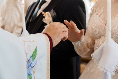 Dokunaklı bir sahnede bir Ortodoks rahibi, düğün törenlerinde bir gelin ve damadın ellerini kutsarken, birlik ve inancı simgelerken, süslenmiş altın işlemeler ve narin beyaz danteller sergilenirken tasvir ediyoruz.