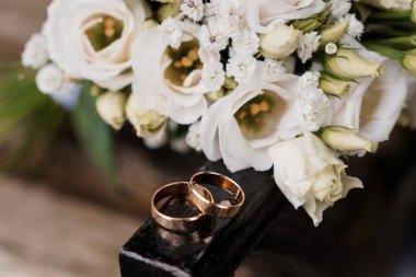 Sarmalanmış altın alyansların yer aldığı romantik yakın plan bir resim. Siyah ahşap bir yüzey üzerinde, narin bir buket beyaz gül ve çiçeğin arasında duruyor.