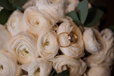 Altın alyansların yakın çekimi beyaz ranunculus çiçeklerinin arasında duruyor, aşkı ve bağlılığı narin, romantik bir tarzda sembolize ediyor.