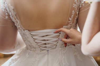 Gelinlerden birinin fildişi gelinliğini bağlarken, çiçek desenleri sergilerken, korse detaylarını anlatırken ve romantik bir hazırlanma sahnesi çekilirken.