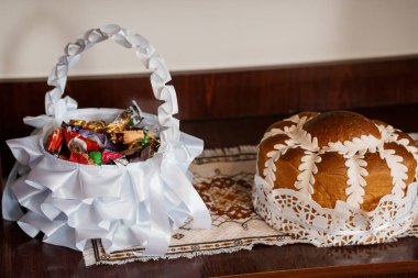 Güzel düzenlenmiş bir natürmortta beyaz dantelli yuvarlak bir Paskalya ekmeği ve koyu ahşap bir yüzey üzerinde sunulan renkli çikolata şekerlerle dolu bir sepet sergileniyor.