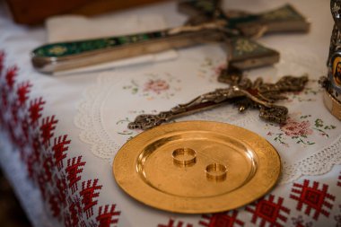 Yakın plan, bir Ortodoks Hıristiyan düğün ritüeli içinde, süslü bir haçın yanında, bir paten üzerinde altın alyansların kutsal alışverişini tasvir eder.