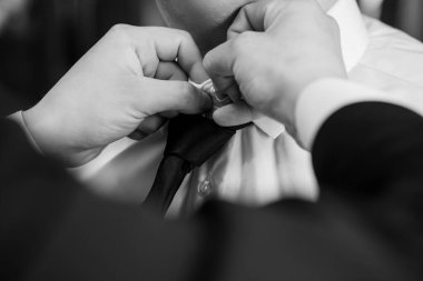 Yakın plan, siyah-beyaz bir fotoğraf bir damadın kravatının düzeltilmesini, düğün hazırlıklarını, samimiyetini ve resmi kıyafetlerini sembolize edişini gösteriyor.