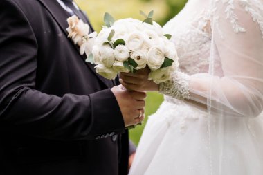 Romantik düğün sahnesinde damadın beyaz bir gül buketini nazikçe tutuşu, gelinlerin boncuklu elbisesi görünür, aşkı, bağlılığı ve klasik bir kutlamayı simgeliyor.