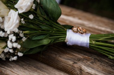 Özel düğün sahnesinde beyaz bir gül buketine bağlanmış altın alyanslar aşk ve bağlılığı simgeliyor. Kırsal, doğal ışıklıklar romantik havayı çağrıştırıyor.