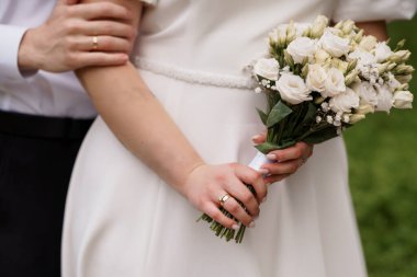 Damat elini beline koyup, sevgiyi, bağlılığı ve kutlamaları sembolize ederken, bir gelinin narin beyaz bir buketi tutuş şeklindeki görüntüsü.