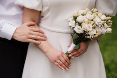 Yakın plan çekimde, bir gelin ve damadın elleri açık hava kutlaması sırasında elinde güzel bir buket beyaz gülle alyansla görüntülenir.