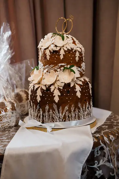 Güzel dekore edilmiş, buğday ve beyaz çiçek aksanıyla süslenmiş iki katlı bir düğün pastası beyaz bir masa örtüsünün üzerinde duruyor. Altın yüzükler bağlılığı simgeler, düğün temaları için mükemmeldir.