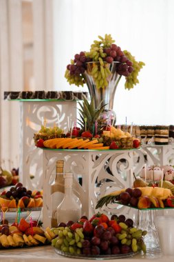 Bolca düzenlenmiş meyve büfeleri üzüm, ananas, çilek ve kavun gibi canlı meyveler süslü kristal vazolarda ve beyaz dekorda sunulmaktadır.