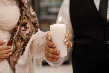 Samimi bir düğün sahnesinde bir gelin ve damadın elinde yanan beyaz sütun mumu, birlikteliği, aşkı ve yeni başlangıçları simgeleyen zarif dantelli bir elbise vardır.