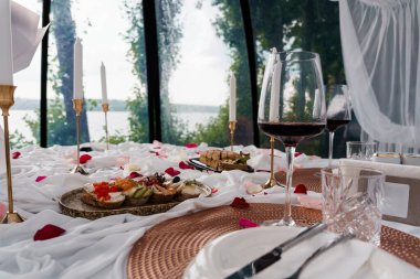 Şarap kadehleri, gül yaprakları ve göl kenarındaki beyaz masa örtüsünün üzerindeki aperatiflerin yer aldığı samimi bir akşam yemeği düzenlemesi romantik bir ortam yaratıyor.