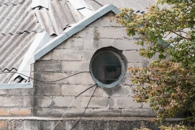 Yıpranmış, metal çatılı gri taş bir duvara açılan dairesel bir pencere ve bir ağaç dalı gölgeli bir iç mekâna göz atmamızı sağlıyor.