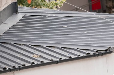 Bu görüntü muhtemelen fırtınadan sonra gri bir binadaki hasarlı metal çatıyı gösteriyor. Görünür virajlar ve kayıp bağlayıcılar nedeniyle onarılması gerekiyor