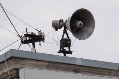 Bir binanın çatısına yerleştirilmiş yıpranmış bir sivil savunma sireni elektrik bileşenleri, gri tonlar ve kentsel çevre detaylarıyla yaşlanan bir iletişim sistemini gözler önüne seriyor.