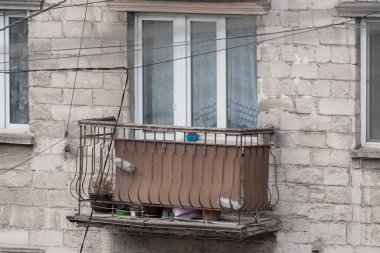 Yıpranmış bir şehir balkonu, paslı metal korkuluklara tünemiş iki güvercin, ağ perdeleri ve beton binanın dış tarafındaki günlük nesnelerin arasında yer alıyor.