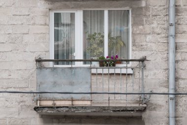 Kırsal bir balkon, yıpranmış beton bir apartmana bağlı pembe çiçekler de dahil olmak üzere canlı saksı bitkilerini beyaz bir pencere çerçevesine sergiliyor.