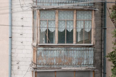 Tahta pencereli yıpranmış bir balkon ve narin dantel perdeler eski bir Avrupa binasında çürüme belirtileri gösteriyor, nostaljik, melankolik bir havayı çağrıştırıyor.