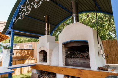 A cozy outdoor kitchen features a stone wood-fired pizza oven integrated with a smoker, sheltered under a blue pergola, showing wood and cooking tools
