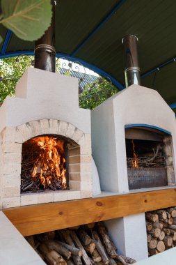 Güzel bir çift katlı tuğlalı pizza fırını, içinde kükreyen bir ateşle, yeşil bir pergola altında, alttaki ahşap depolama bölümü ile birlikte resmediliyor.