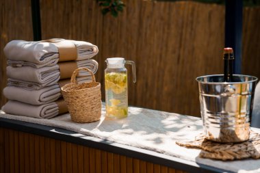 Düzgünce istiflenmiş keten havluları, ferahlatıcı bir sürahi limon suyu, buz kovasında bir şişe şarap ve bir sepet, yaz yaşam tarzı tasvirleri için ideal.