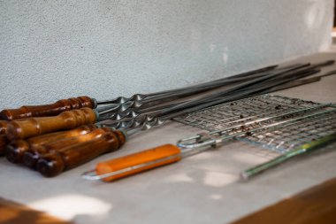 Yakın plan paslanmaz çelik ızgara ızgara ızgaraları ve ahşap saplı şişler dokulu gri bir taş tezgahın üzerinde duruyor. Açık havada et pişirmek için mutfak aletleri.