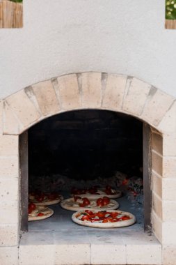 Geleneksel bir taştan pizza fırını içeride birkaç margherita pizzası pişirir ve açık mutfak ortamında sıcak ışıklandırmalı İtalyan yemekleri sergiler.