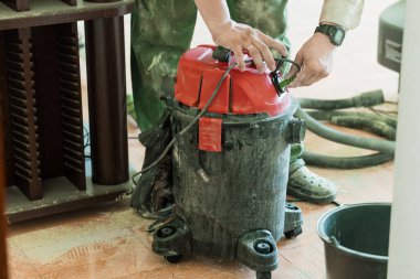 Koruyucu giysiler giyen bir ressam, atölye zeminindeki talaşları temizlemek için, ahşap mobilya ve aletlerin etrafındaki döküntülere odaklanmak için kırmızı bir süpürge kullanır.