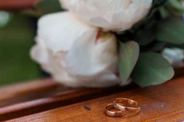 Aşkı, bağlılığı ve evliliği sembolize eden beyaz şakayık demetinin yanında, ahşap bir yüzeyde dinlenen altın alyansların yakın çekimi.