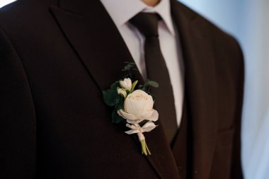 Siyah smokin giyen bir damadın yakın plan fotoğrafı, üzerinde okaliptüs yapraklı ve ipek kurdeleli, narin bir gül yaka çiçeği ve somut bir düğün modayla birlikte.