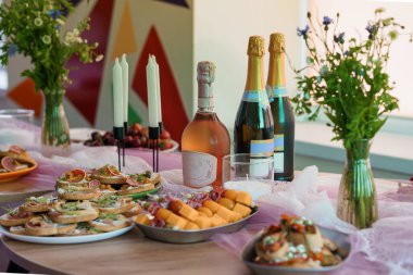 Şatafatlı şampanya şişeleri, çeşitli aperatifler, taze meyve ve renkli çiçek aranjmanları kutlama kahvaltısı için pembe kaplı bir masada sergileniyor.