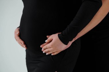 Hamile bir kadının elleri, dar, siyah bir kurdeleli kıyafet giyerken yuvarlak göbeğini sallarken, annelik beklentisinin hassas bir anını resmediyordu.