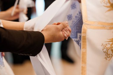 Yakın plan çekimde, bir düğün ya da dini tören sırasında el ele tutuşan bir damat, bir rahibin rehberliğinde, altın işlemeli beyaz elbiseler altında, inanç ve birliği sembolize eden bir gelinin elini tutuyor.