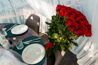 Güzel hazırlanmış romantik bir yemek sahnesinde iki kişilik zarif bir masanın yanında büyük bir buket kırmızı gül yer alır. Aşk ve kutlama duyguları uyandırır.