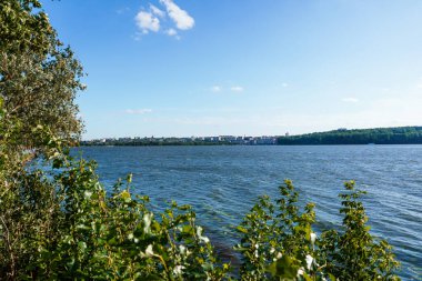 Açık mavi gökyüzünün altında yeşil ağaçların arasına kurulmuş uzak bir kasaba silueti ile Hudson Nehri 'nin yer aldığı canlı, geniş açılı bir manzara, huzurlu su yansımasını vurguluyor.
