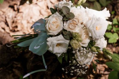 Beyaz güllerden ve narin bebek nefesinden oluşan güzel bir düğün buketi. Çiçekler arasında duran zarif altın alyanslar ve doğal bir ormanda geçen mavi kurdele.