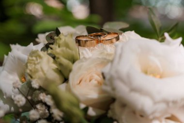 Gül sarısı alyansların yakın plan fotoğrafı bir buket fildişi beyaz gülün üzerinde özenle duruyor. Aşkı, evliliği ve bağlılığı simgeleyen yumuşak, romantik bir estetik.