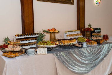 Bir etkinlik ortamında geniş bir dizi hamur işi, taze meyve düzenlemeleri ve gümüş pullu masa örtüsü içeren lüks bir tatlı büfesi, düğünler için ideal.