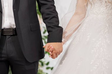 Siyah takım elbiseli bir damadın resmi bir tören sırasında beyaz gelinlik giyen bir gelinle el ele tutuşması, aşkı ve bağlılığı simgeliyor.