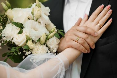 Altın alyansları sergileyen bir düğün çifti, güzel beyaz bir çiçek buketiyle sarmalanmış. Sevgi ve bağlılığı sembolize eden neşeli romantik jest